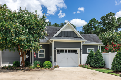 Single-story gray house with white trim, a single-car garage, landscaped yard, and trees under a blue sky with scattered clouds.