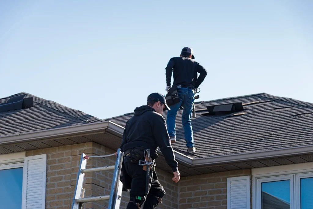 Two workers in safety gear are on the roof of a brick house, one standing on the roof and the other climbing a ladder, both appearing to inspect or repair the shingles.